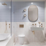 Light blue and white marble-look tiled bathroom with shower over bath, white wall-hung basin, pill-shaped mirror and gold fittings