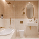 Peach and cream marble-look tiled bathroom with shower over bath, white wall-hung basin, pill-shaped mirror and gold fittings